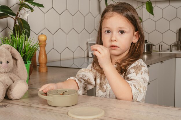 Little dark haired girl 34 years old taking piece of food out from pastel gray silicone snack cup in kitchen Toy bunny