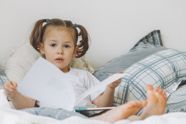 Little darkhaired girl  with two ponytails sits on bed with pillows and reads book