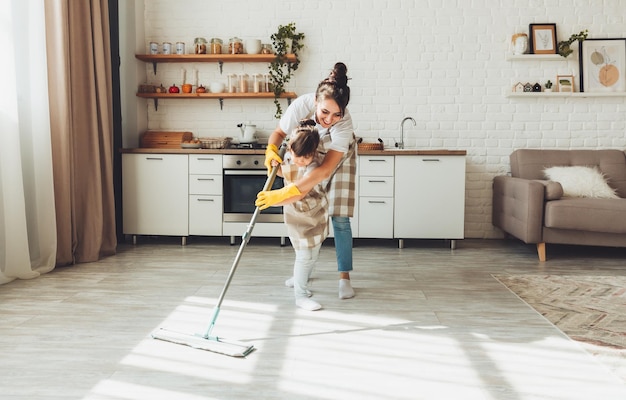 小さな娘と彼女のお母さんが家を掃除します 子供が台所の床を洗います かわいいヘルパーの女の子がモップで床を掃除します 幸せな家族が部屋を掃除します