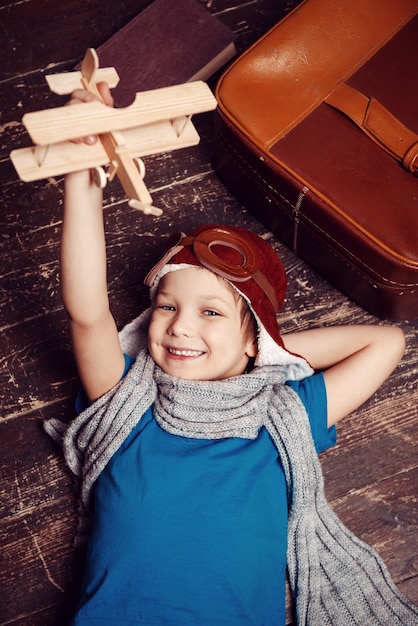 Little day dreamer. Top view of happy little boy in pilot headwear and eyeglasses lying on the hardwood floor and playing with wooden planer
