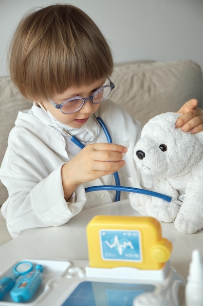 청진기가 있는 의료 코트를 입은 작은 의사가 테디 베어 장난감에 눈 치료를 합니다.