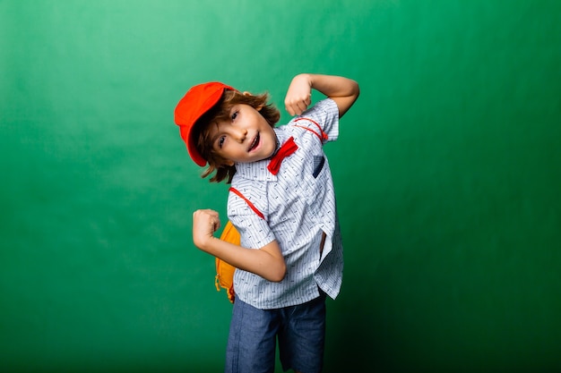 Little emotional 7-year-old boy in a red cap and with a yellow backpack. Standing on a green background, having fun.