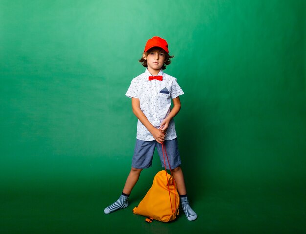 Little emotional 7-year-old boy in a red cap and with a yellow backpack. Standing on a green background, having fun.