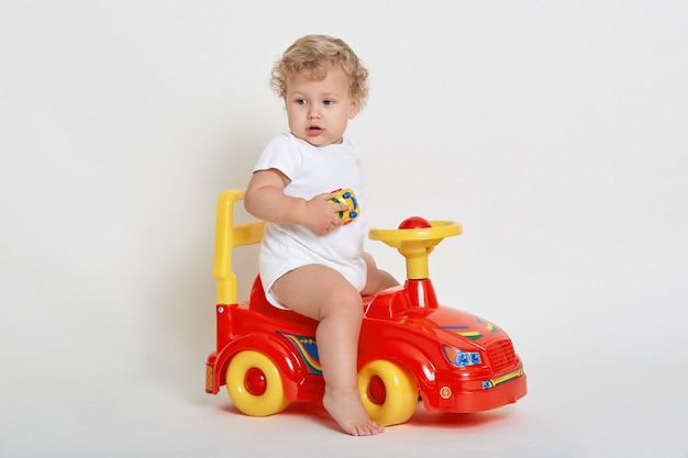 Little European boy riding tolocar barefoot, dresses white suit, blonde infant with curly hair looking away