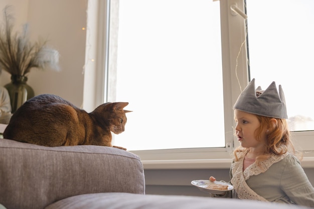 Little fairskinned redhaired girl looks at cat holds plate of cookies standing by window during day Child concept