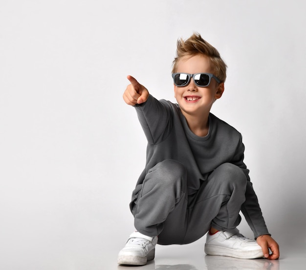 Photo little fashion boy with happy toothy smile on face wearing sunglasses and trendy casual sportswear squatting pointing finger forward fulllength studio shot over white background
