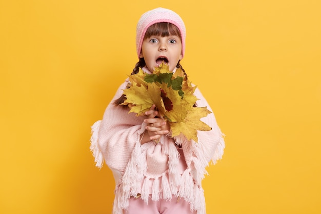 Little fashionable girl in warm clothes holding bouquet of autumn leaves