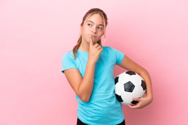 분홍색 배경에 격리된 어린 축구 선수 소녀는 올려다보는 동안 의심을 품고 있습니다