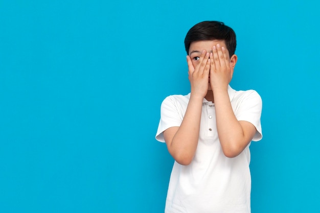 Фото Маленький испуганный азиатский мальчик в белой футболке закрывает лицо руками и выглядывает