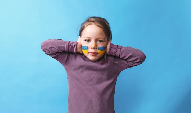 A little frightened girl with a Ukrainian flag painted on her cheek covers her ears with her hands so as not to hear anything on an isolated blue background