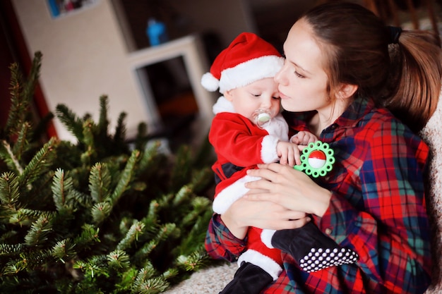 Little funny baby dressed as Santa Claus and young beautiful mother next to the Christmas tree. 