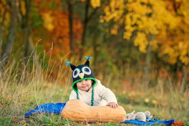 Little funny boy in a knitted cap shows tongue, sitting on grass with pumpkin and autumn trees