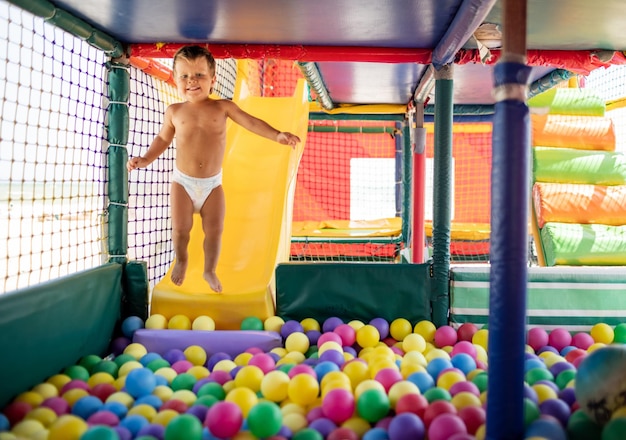 A little funny carefree kid plays in a small playroom and rolls from a bright little slide into a pool with small colorful balls