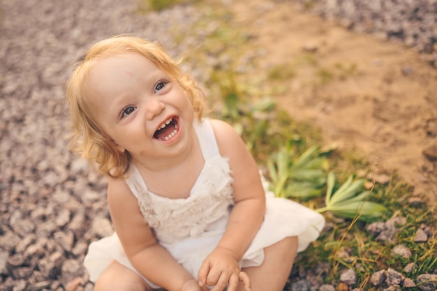 Маленькая забавная милая блондинка девочка-малышка с кудрями в белом платье и с грязью на лице сидит