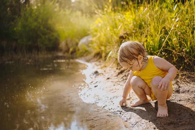 Little funny cute blonde girl child toddler in yellow wet bodysuit playing by the lake waterside