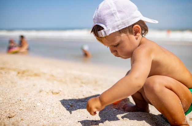작고 재미있는 아이는 밝은 휴가에 뜨거운 여름 태양 아래 모래 바닥에 잔잔한 푸른 바다에서 조개와 자갈을 수집합니다