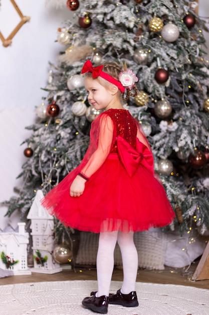 Little funny girl in a red dress stands near the Christmas tree