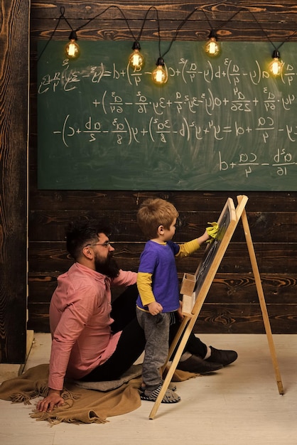数学を学ぶ小さな天才。黒板で方程式を解くために子供を助ける教師または父親。床に座っているピンクのシャツのひげを生やした男