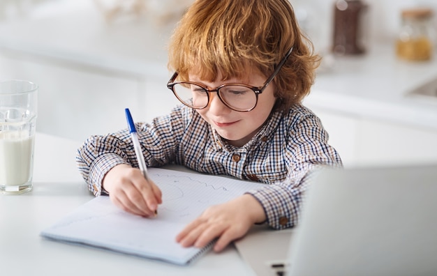 少し天才。しつこい活気のある生姜の子供は、大きな眼鏡をかけ、白いテーブルに座って、自分の考えを注意深く書き留めます