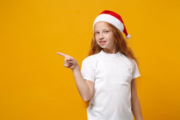 Little ginger kid Santa girl 12-13 years old in white t-shirt Christmas hat isolated on yellow background. New Year 2020 celebration holiday concept. Mock up copy space. Pointing index finger aside.