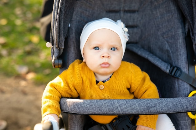 A little girl 1 year old dressed in a yellow shirt and a white hat sits in a grey stroller in the park