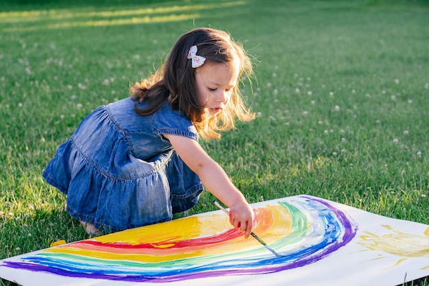 2-4세 어린 소녀는 녹색 잔디에 앉아 큰 종이에 무지개와 태양을 그립니다.