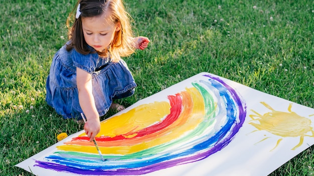 2〜4歳の少女は、緑の芝生の上に座って、大きな紙に虹と太陽を描きます