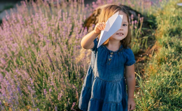 Маленькая девочка 3-4 лет с темными волосами в джинсовом платье на солнце запускает бумажный самолетик среди больших кустов сирени лаванды