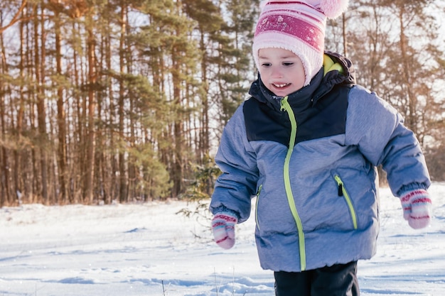 Little girl 34 years old in winter clothes overalls hat and mittens runs on snow and laughs against pines and fir