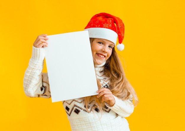 A little girl of 8 years old with blond hair in a Santa Claus hat holds a white sheet of paper on a yellow background.