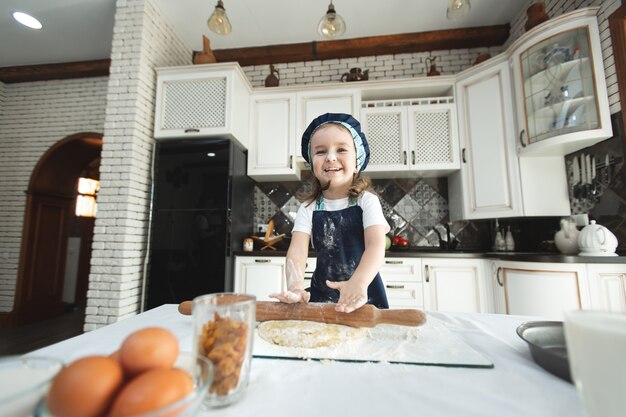 A little girl in an apron and a chef's hat is rolling out cookie dough, laughing.