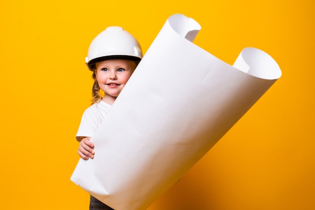 Little girl architect in the construction helmet with a poster isolated on yellow wall