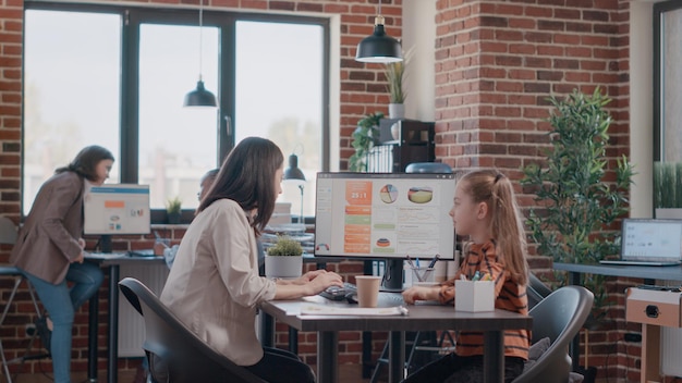 Little girl arriving back from school at single mom workplace. Working mother greeting child at office while working on business on computer for project planning and strategy. Employed parent