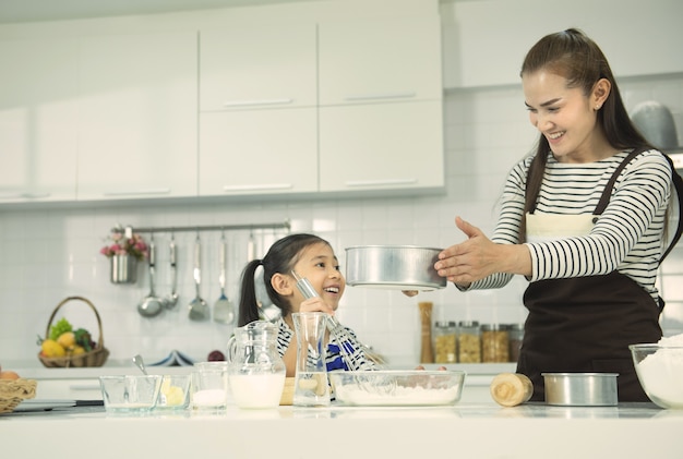 Foto bambina e mamma asiatica in grembiuli che giocano e che ridono mentre impastano la pasta in cucina. pasticceria artigianale.