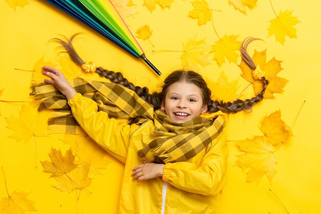 단풍잎이 떨어진 가을 노란색 배경의 어린 소녀는 화려한 우산으로 거짓말을 하고 미소를 짓고 그 아이는 밝은 가을 분위기로 비옷과 스카프를 입고 누워 있습니다