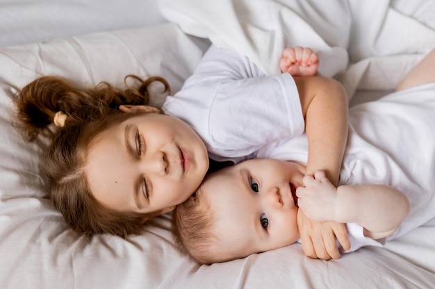 Bambina e bambino in abiti bianchi sono sdraiati a letto su lenzuola bianche. fratello e sorella si crogiolano a letto. sonno e riposo dei bambini. hygge, stile di vita. spazio per il testo. foto di alta qualità