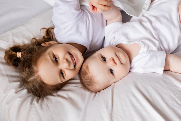 Little girl and baby in white clothes are lying in bed on white bed linen brother and sister