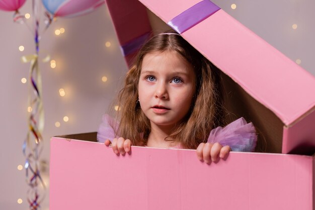 Little girl in a beautiful dress is hiding in a huge pink cardboard gift box happy birthday