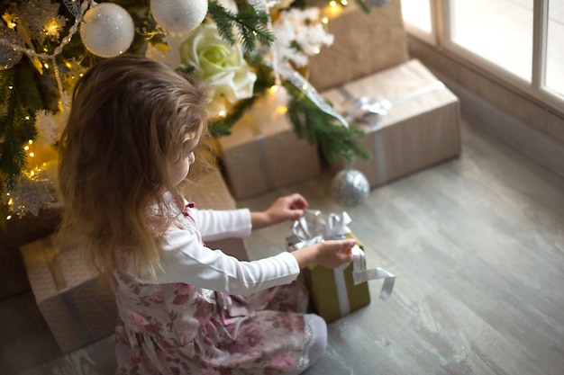 little girl in beautiful dress is sitting under christmas tree with gift box and bow. light from a large window, dark room, home comfort, christmas, magic and joy. 