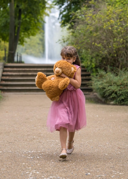 A little girl in a beautiful pink dress runs quickly and holds a large teddy bear in her hands