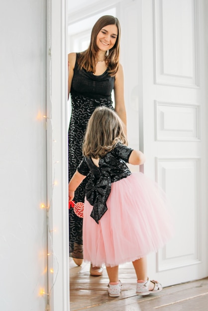 Little girl in beautiful pink dress with tutu skirt looks at the door with her mother