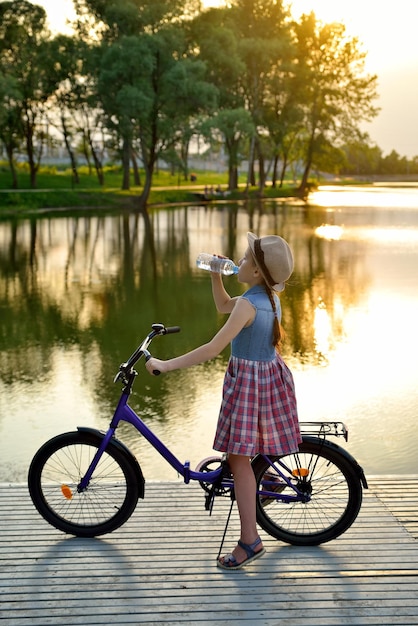 자전거를 탄 어린 소녀는 강둑의 일몰에 서서 플라스틱 병의 물을 마신다
