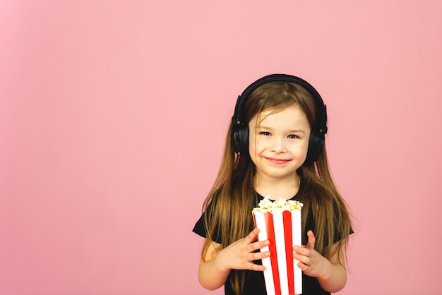 Little girl in big headphones watches a movie, eats pop corn and smiles on a pink pastel background