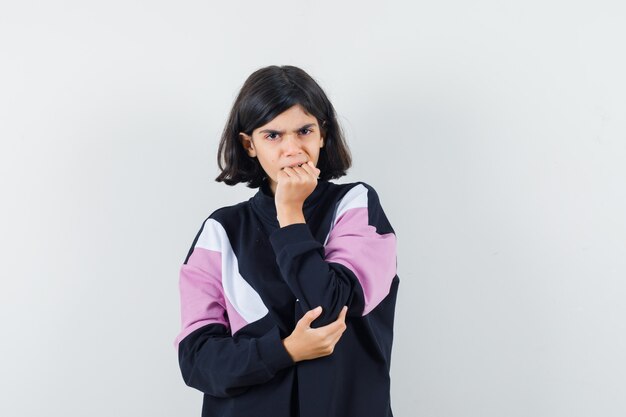 Little girl biting her fist emotionally in shirt and looking frightened , front view.