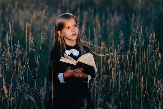 A little girl in a black dress stands in tall grass and holds a green book in the light of the setting sun. a child with a pensive facial expression.