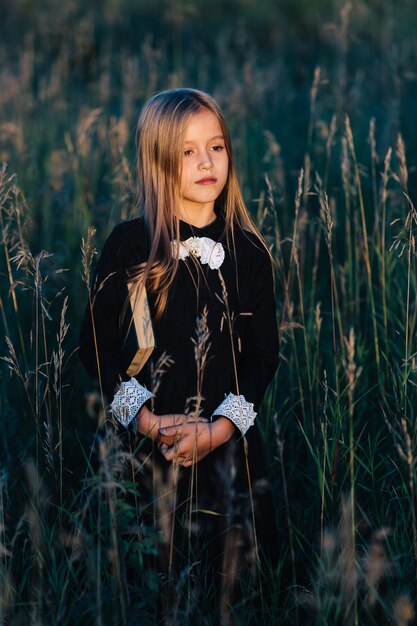 A little girl in a black dress stands in tall grass and holds a green book while looking at the sunset.