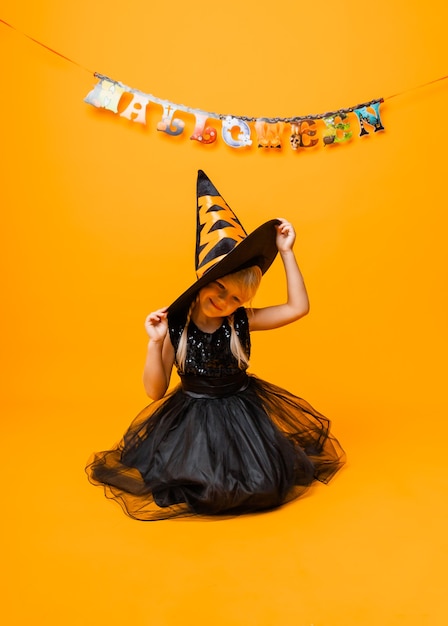 Маленькая девочка в черном костюме Хэллоуина смеется и смотрит в камеру, прыгает и веселится, изолированные на желтом фоне. Хэллоуин