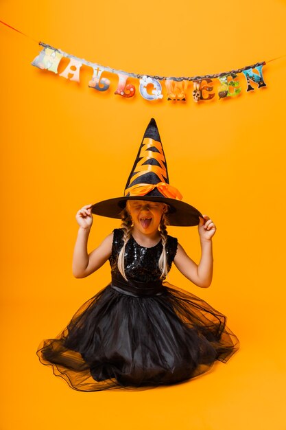 Foto bambina in costume di halloween nero che ride e guarda la telecamera, salta e si diverte, isolata su sfondo giallo. halloween