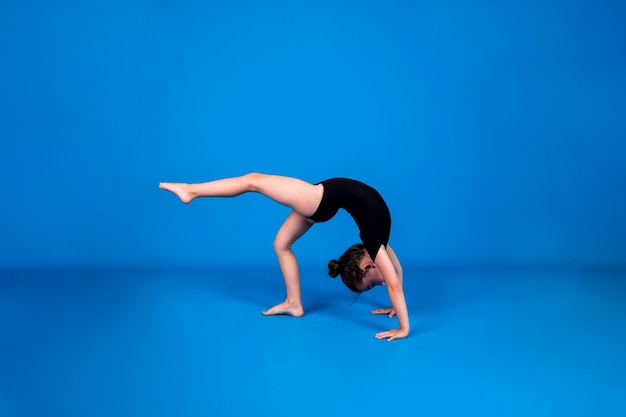 A little girl in a black swimsuit performs a rhythmic gymnastics exercise on a blue background with a place for text