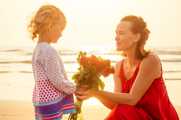 小さな女の子のブロンドは、ビーチで母親にバラの花束を贈ります海の幸福な母の日までに花を持つ美しい母と娘バレンタインデー2月14日